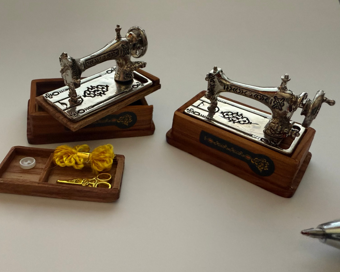 Two miniature sewing machine models on wooden bases with a small tray containing yellow thread and scissors.