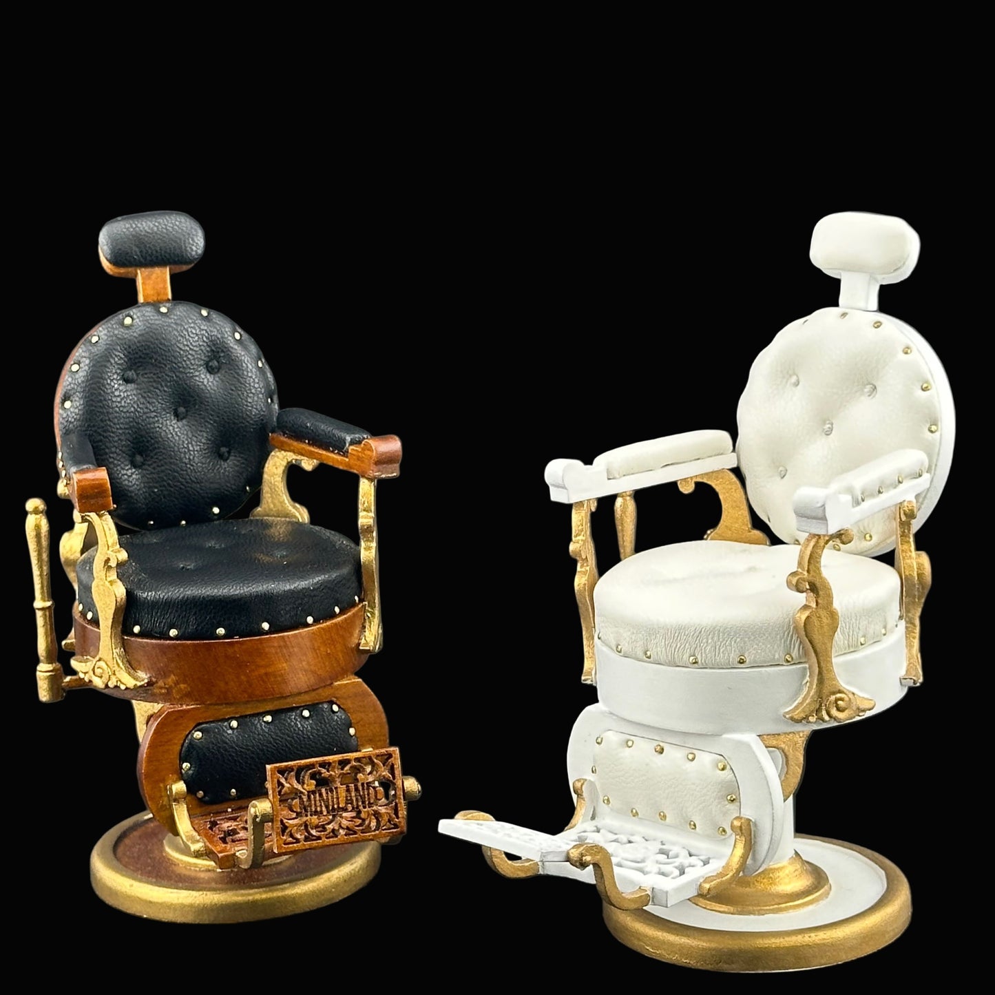Two miniature barber chairs, one black and gold and one white with gold accents, on a dark background.