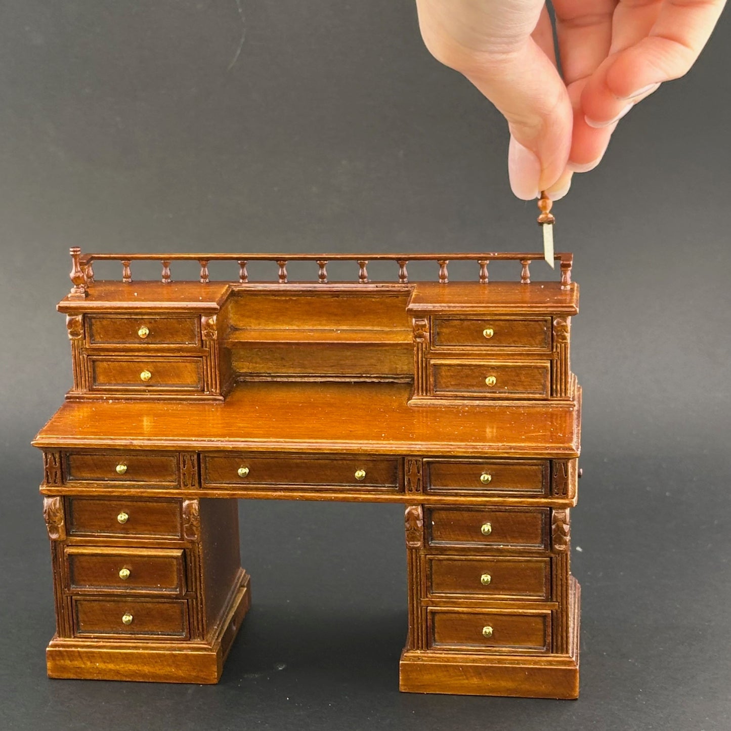 Miniature wooden desk with drawers and a hand holding a small object on top against a dark background